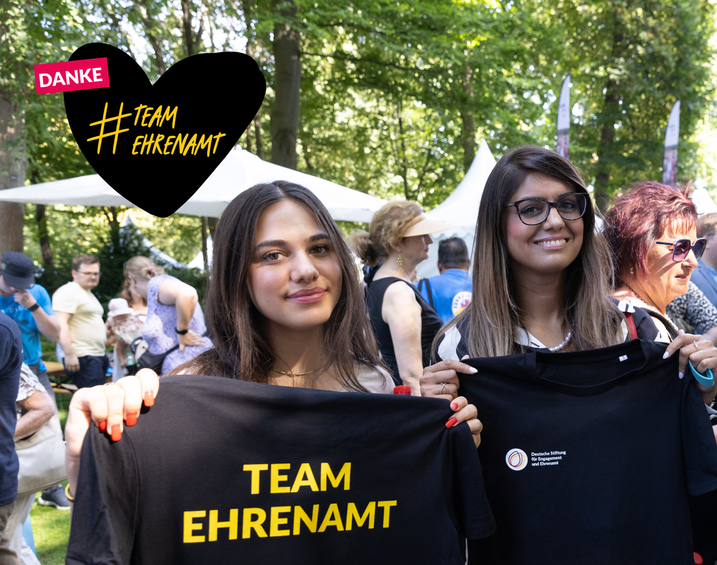 Foto von zwei jungen Frauen, die T-Shirts mit dem Logo der DSEE und dem Schriftzug Team Ehrenamt in die Hände halten. Gezeichnetes Herz mit dem Text Danke #TeamEhrenamt