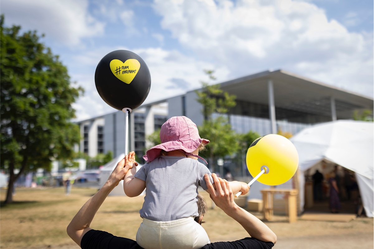 Foto von einem Kind auf den Schultern einer erwachsenen Person mit zwei Luftballons. Darauf ein Herz mit dem Text #TeamEhrenamt
