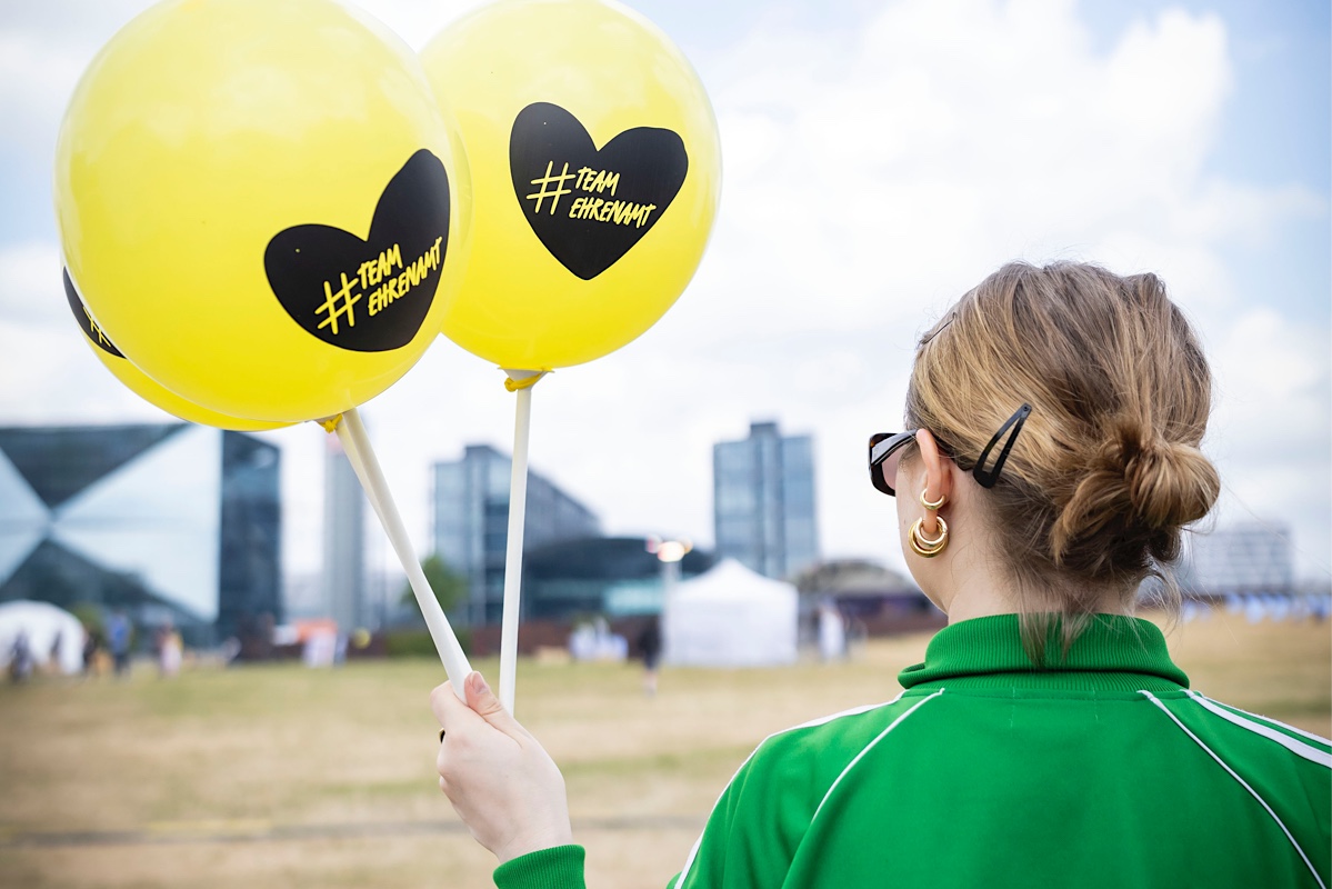 Foto von einer jungen Person mit zwei Luftballons. Darauf ein Herz mit dem Text #TeamEhrenamt