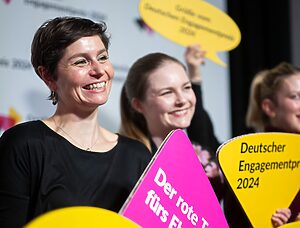 Zwei weiblich gelesene, lächelnde Frauen, die ein magentafarbenes Schild hochhalten, auf welchem "Der rote Teppich fürs Ehrenamt" steht