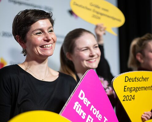 Zwei weiblich gelesene, lächelnde Frauen, die ein magentafarbenes Schild hochhalten, auf welchem "Der rote Teppich fürs Ehrenamt" steht