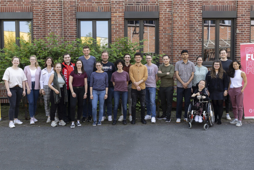 Gruppenbild mit 20 jungen, lächelnden Erwachsenen vor einem Backsteingebäude