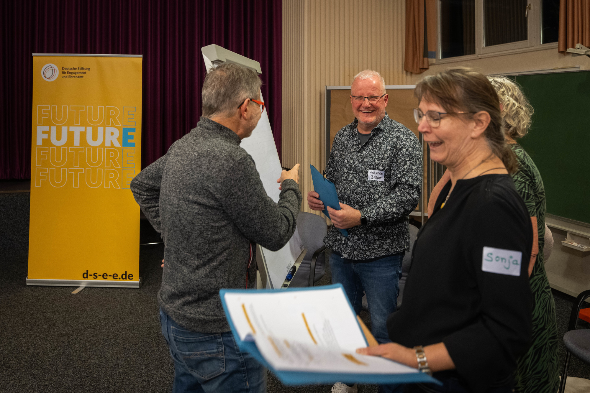 Bild mit zwei älteren Männern und zwei älteren Frauen in einer Workshop-Diskussion. Sie lachen und stehen um ein Flipchart. Im Hintergrund steht ein Rollup-Aufsteller mit Text: "Deutsche Stiftung Engagement und Ehrenamt, d-s-e-e.de , Futur E